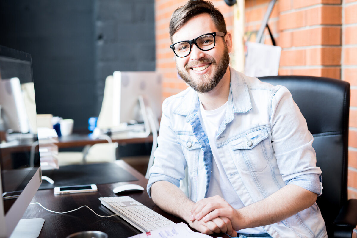 reviews business manager wearing casual clothes smiling happily at camera while sitting at computer desk
