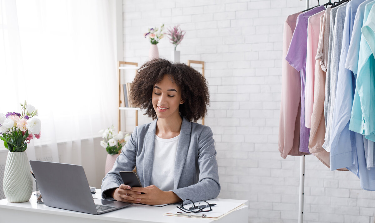 Great reviews from customers to small business of fashion store. Smiling african american woman reading in smartphone, sitting at table with laptop in living room interior with rack with clothes