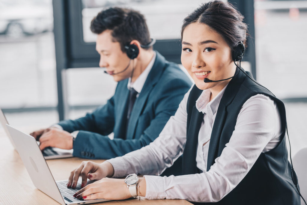 young woman in headset smiling at camera while working with male colleague in call center