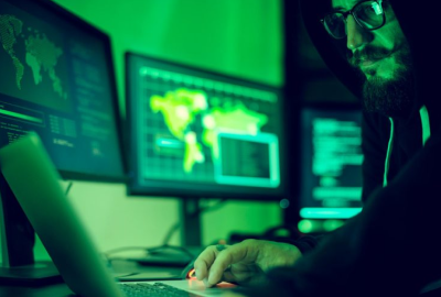 A man in a hoodie is working on a laptop in front of two monitors.